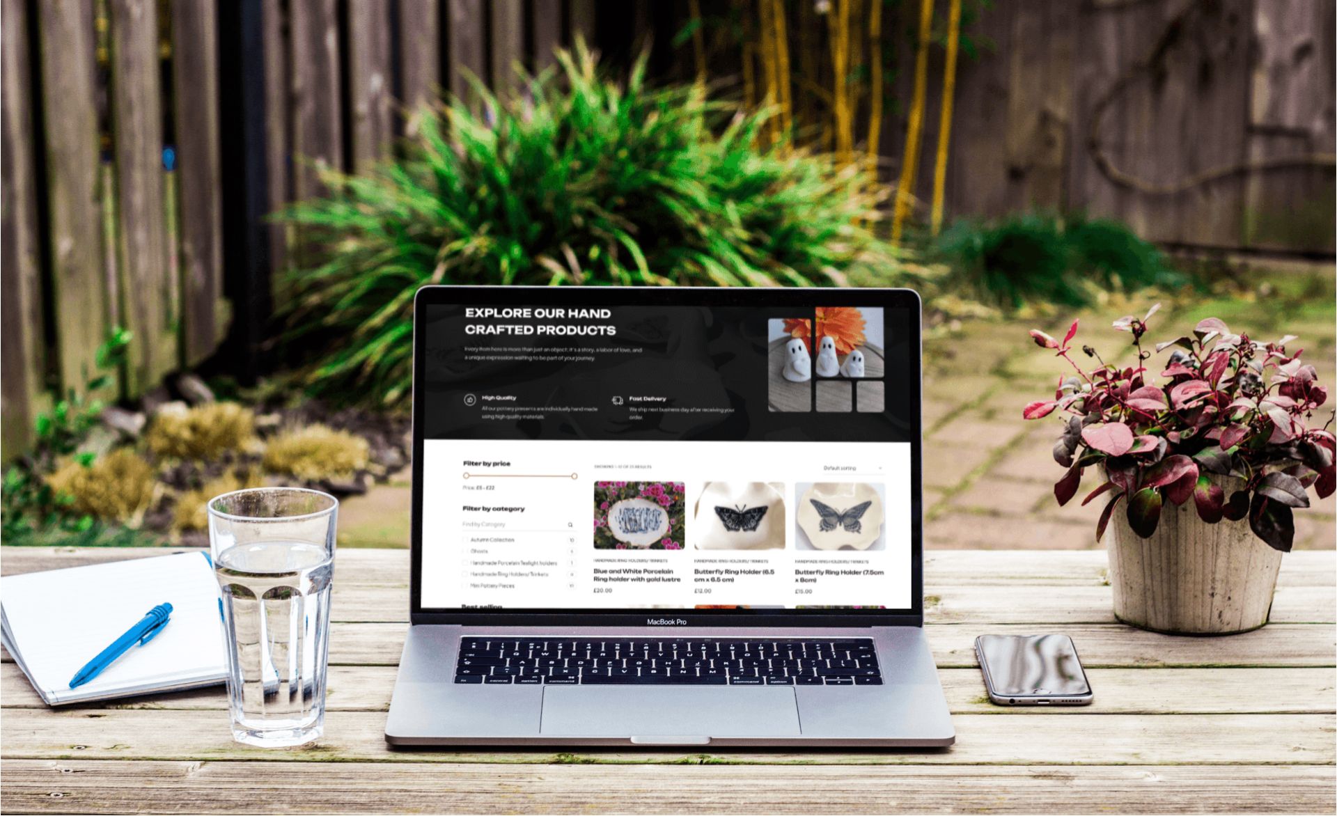 A laptop displaying a website is set on a wooden table outdoors, alongside a glass of water, a notepad, and a smartphone. There's a potted plant nearby, and a garden is visible in the background.