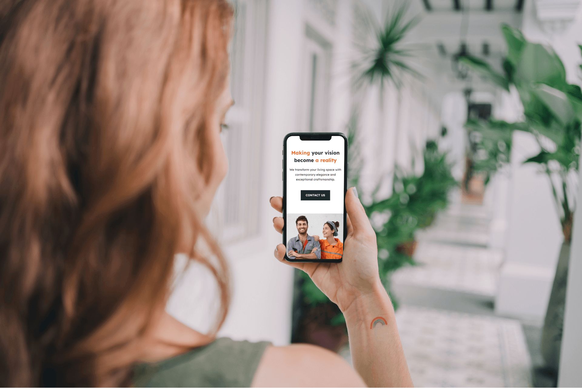 A person with long hair holds a smartphone displaying a webpage by stockport web design company iindigo with text and a photo of two people. They stand in a corridor with plants and patterned flooring.
