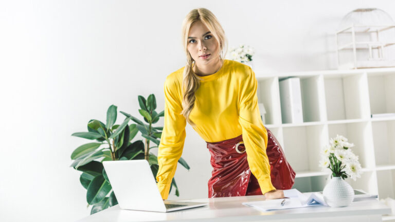 A woman in a yellow shirt is sitting at a desk with a laptop.